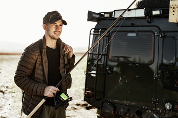 Young fisherman standing near his car and holding fishing equipment