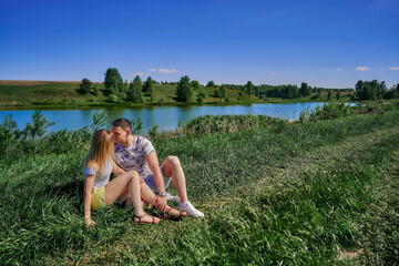 A couple in love are kissing while sitting on the grass by the river on a sunny summer.