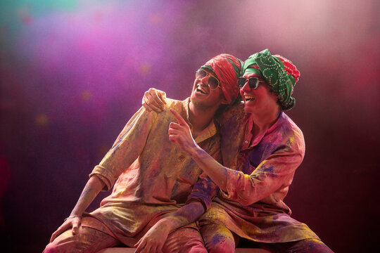 A HAPPY YOUNG MEN SITTING WITH FRIEND LOOKING AWAY AND POINTING WITH GULAL ALL OVER THEM	