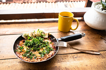 Szakszuka, jajka smażone w pomidorach na patelni, pyszne śniadanie na stole z tropikalnym widokiem w tle. © insomniafoto