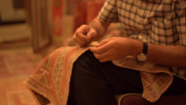 Guy master crafts small designed carpet with traditional local ornaments sitting in local shop against blurred car in evening close view.