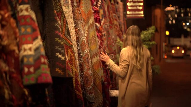 Young woman tourist with long loose fair hair touches coloured carpet with traditional ornaments and walks along street in evening.