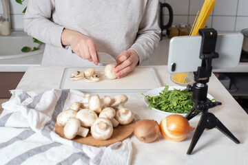 Woman food blogger in her home kitchen cut mushrooms for cooking vegetarian pasta. Live stream with subscribers, making video lesson with phone. Distance online learning. Cooking courses tutorials