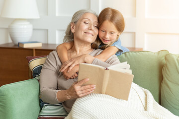 Loving grandmother teaching granddaughter reading book sitting on sofa. Kid girl embracing grandma or baby sitter during telling fairy tale, nanny granny spending time with preschool grandchild.