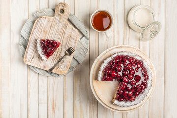 Cherry pie with a cut piece on a white wooden board on a light background with a cup of tea and a sugar bowl.