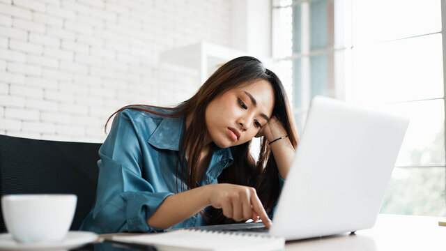 Portrait of bored young asian woman typing laptop in office. Freelance employees sleeping lying head on hand on computer desk feel boring. Lifestyle of routine employees concept.
