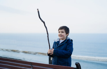 Outdoor portrait Active kid playing with wooden stick with blurry blue ocean seaside background, Happy boy having fun playing by the sea on Summer, Healthy Child relaxing outside in sunny day Spring.