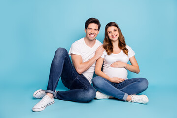 Photo of optimistic pregnant couple sit hug wear white t-shirts jeans sneakers isolated on cyan color background