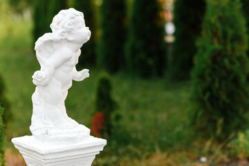 statue of baby angel with wings against green garden. The cute little cupid angel sculpture statue...