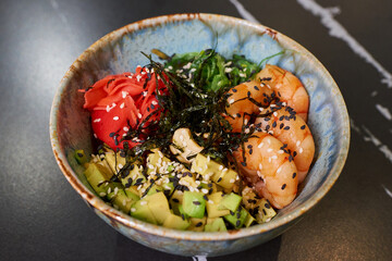 A close-up photo of a poke bowl with Chuka Wakame seaweed, wasabi, avocado, cashew, nori, and shrimps on a black stone table.