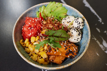 A top view of a poke bowl with tuna, avocado slices, wasabi, corn, peanut, sesame, and Philadelphia cheese on a black stone table.