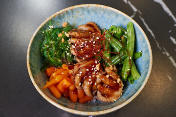 A top view of a poke bowl with octopuses, Wakame seaweed, carrot, peanut, sesame, white rice on a black stone table.