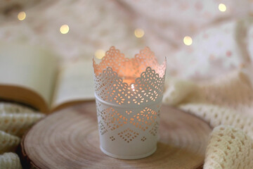 Candle holder with lit candle, wooden tray, open book and knitted blanket at home. Selective focus. 