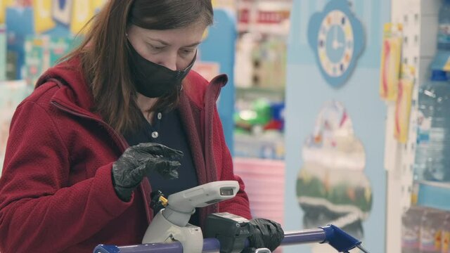 Woman in mask and gloves against virus makes contactless purchase goods in store. She introduce product code into electronic validator. Concept of safe ones buy goods during epidemics, pandemics