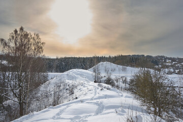 Fototapeta na wymiar Snowdrifts on winter snow covered small hills, trees and sun shine glow in yellow-blue sky.