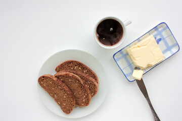 Cup of black herbal tea, slices of dark rye and whole wheat bread with sesame seeds and butter on a plate on white table background. Healthy breakfast concept. Top view, copy space 