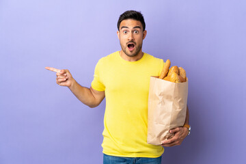 Young caucasian man holding a bag full of breads isolated on purple background surprised and pointing finger to the side