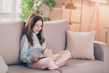 Portrait of attractive cheerful focused preteen girl sitting on divan resting reading book at house...