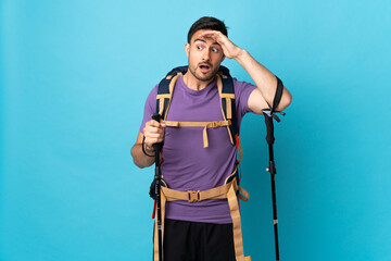 Young caucasian man with backpack and trekking poles isolated on blue background doing surprise gesture while looking to the side
