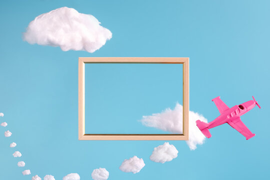 Wooden frame on blue background with abstract shape of clouds and pink airplane with clouds trail. Minimal border composition for your design. Copy space for text