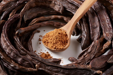 In the foreground, wooden spoon with carob flour. Around and in the background, carob pods. All on...