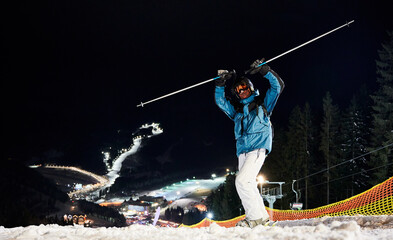 Skier keeping his hands up, standing on hill getting ready for a night ride. Beautiful illuminated ski pistes are down on background in night time. Concept of extreme sport