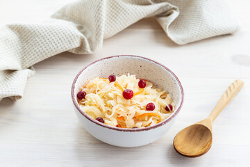 Pickled cabbage with cranberries with a wooden spoon in a bowl on a light table.