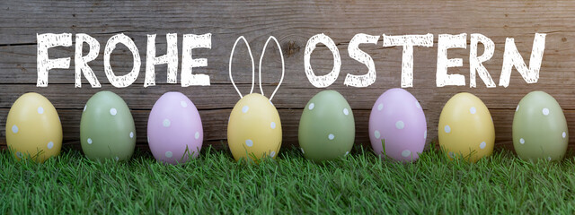 Frohe Ostern Hintergrund Banner Panorama Grußkarte - Viele pastellfarbene gepunktete Ostereier und Hasenohren auf frischer grüner Wiese Gräsern und rustikaler Holzwand Holz Textur