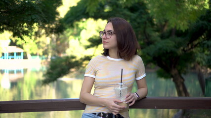 A young woman with glasses stands on a bridge in the park closeup. In the background there is a lake.