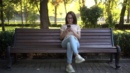 A young woman in glasses sits on a bench with a phone in her hands. In the background nature.