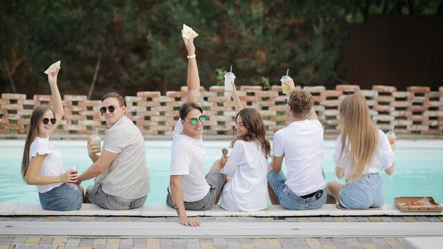 Back view of friends clinking glasses with cocktails sitting by swimming pool on sunny summer day. People have fun at poolside party toasting, drinking beverages at luxury tropical villa