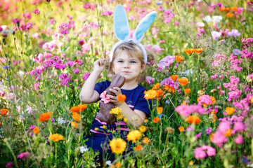 Portriat of adorable, charming toddler girl with Easter bunny ears eating chocolate bunny figure in...