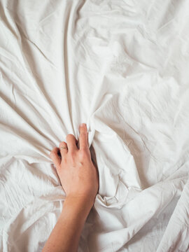 Woman's hand on background of crumpled bed sheet. Creased unmade bed. Woman squeezes white linen. Cozy imperfect house. Non-ideal cozy home.