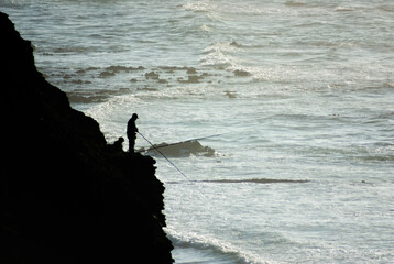 Two fishermen fishing from a cliff in the Ocean. Backlight shot, two silhouettes in the wilderness.