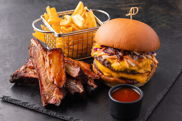 Big yummy burger with double cutlet, fries and fried ribs with barbecue sauce on a black slate...