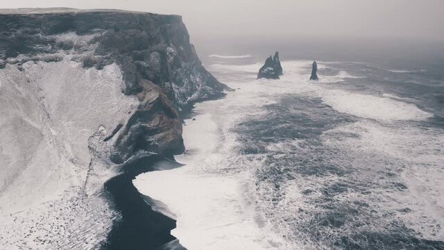 Reynisfjara black sand beach in Iceland with basalt rocks covered with snow aerial view on a stormy winter day. Unreal and spectacular travel destination.