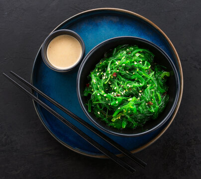 Healthy Wakame Seaweed Salad In A Black Bowl, Peanut Sauce On Side And Chopsticks On A Beautiful Blue Plate Against A Black Slate Background. View From Above.