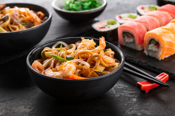 Traditional Asian food set. Wok noodles with shrimps and vegetables in a black bowl, sushi roll set...
