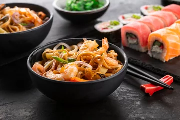 Fototapeten Traditional Asian food set. Wok noodles with shrimps and vegetables in a black bowl, sushi roll set and seaweed salad on a black textured background, angle view. © Goncharuk film