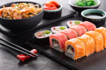 Sushi rolls set, wok noodles and chopsticks side view on black slate board, angle view. Traditional Asian food.