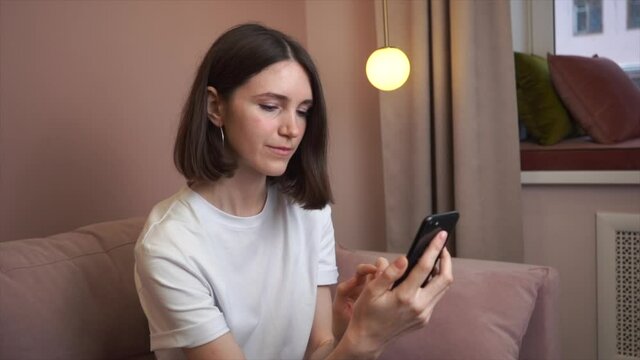 Relaxed young woman using smartphone browsing social networks, playing mobile games, texting while sitting on the couch.Millennial woman spends time at home with mobile gadgets.Side view. Slow motion