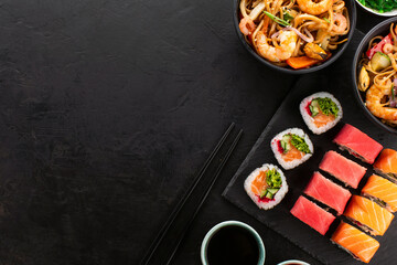 Traditional Asian food set. Wok noodles with shrimps and vegetables in a black bowl, sushi roll and seaweed salad on a black slate background, top view, copy space.