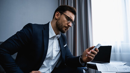Businessman using smartphone near laptop and papers in hotel