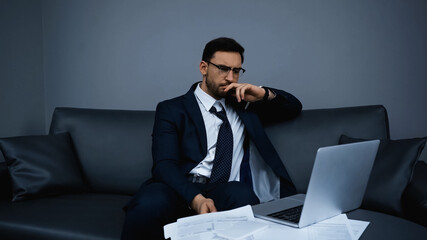 Worried businessman looking at laptop near papers and smartphone in hotel room