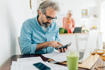 Senior man calculating bills to pay while senior woman cooking in kitchen