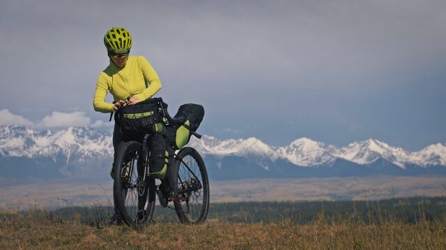 The woman travel on mixed terrain cycle touring with bikepacking. The traveler journey with bicycle bags. Sport tourism bikepacking, bike, sportswear in green black colors. Mountain snow capped.