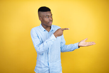 Young african american man wearing a casual shirt standing over yellow background surprised, showing and pointing something that is on his hand