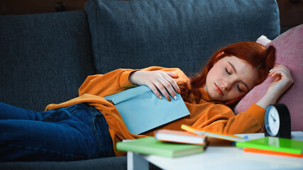 Red haired girl sleeping near book and alarm clock on blurred foreground