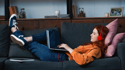 Side view of girl in headphones using laptop near digital tablet on couch