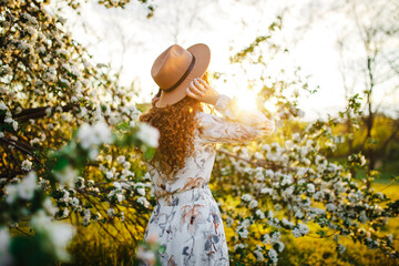 Backview of a young woman among the white blossom of the apple trees at the spring park. Female wearing white dress and beige hat feeling fresh and enjoys good weather on the sunset.